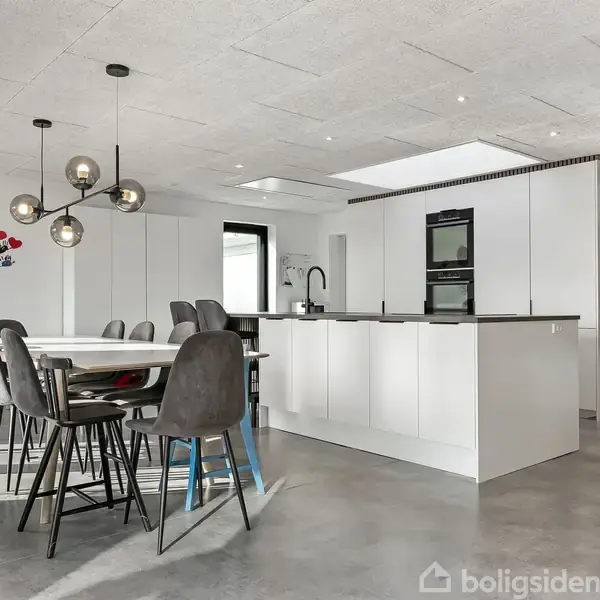Moderne spisebord omgivet af polstrede stole står i et lyst køkken med hvidt interiør, indbygningsovn og lyse lamper. Grønne planter dekorerer væggen, og gulvet er i beton.