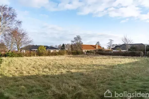 En åben græsmark omgivet af huse ses i et villakvarter med skyet himmel i baggrunden. Træer og buske omkranser området.