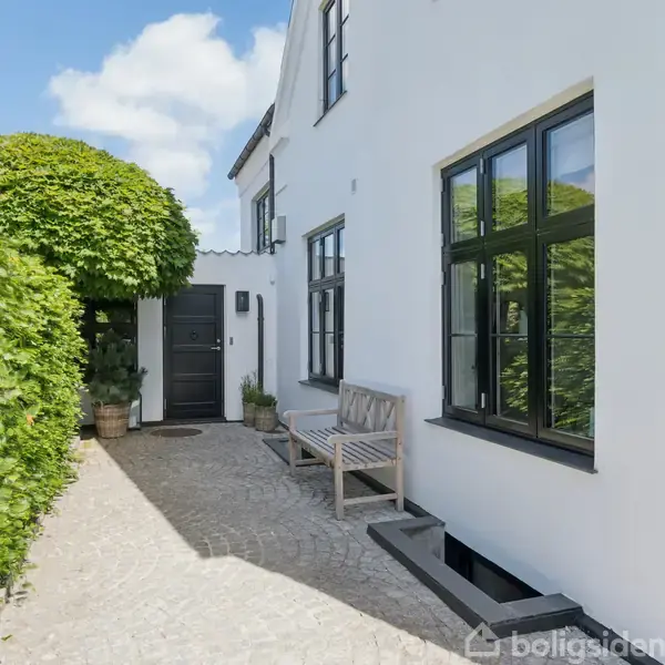 Hvid husfacade med sorte vinduer og dør. Bænken står ved hovedindgangen. Omringet af brostensbelægning og grønne planter i potter under en blå himmel.