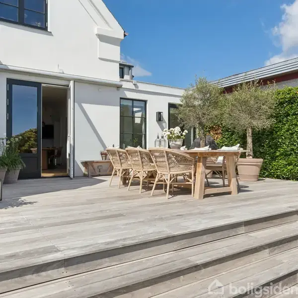 Terrasse med træbord og stole. Bordet er pyntet med planter, omgivet af store plantekrukker og oliventræer. Bagved ses et moderne hus med åbne glasdøre og en grøn hæk.