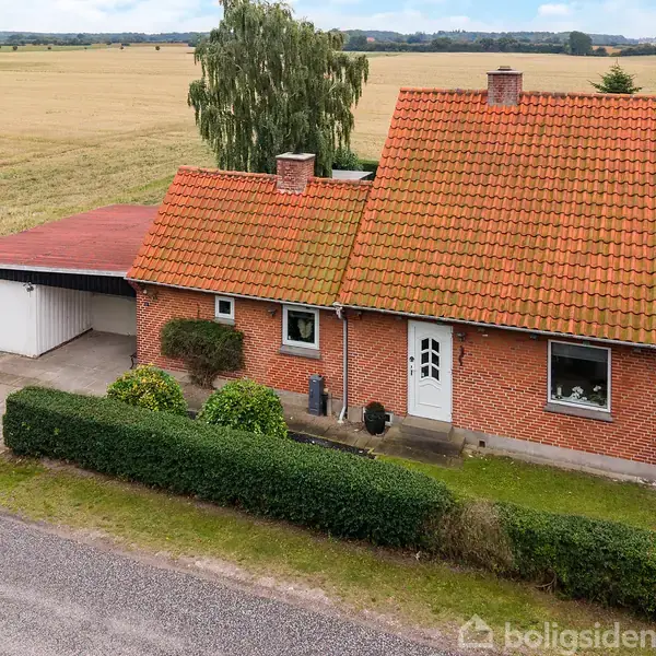 En murstenshus med rødt tegltag og hvid dør står stille på en velplejet grund. Ved siden af er en garage med fladt tag. Omgivelserne er åbne marker.