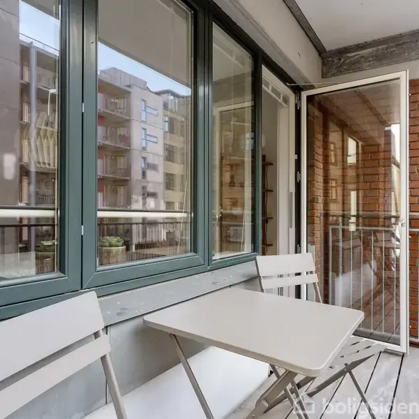 Et sæt bænke og et bord står på en balkon. Balkonen har metalrækværk, og en murstensvæg ses i baggrunden. Vinduer og glasdør giver udsyn til nabobygninger.