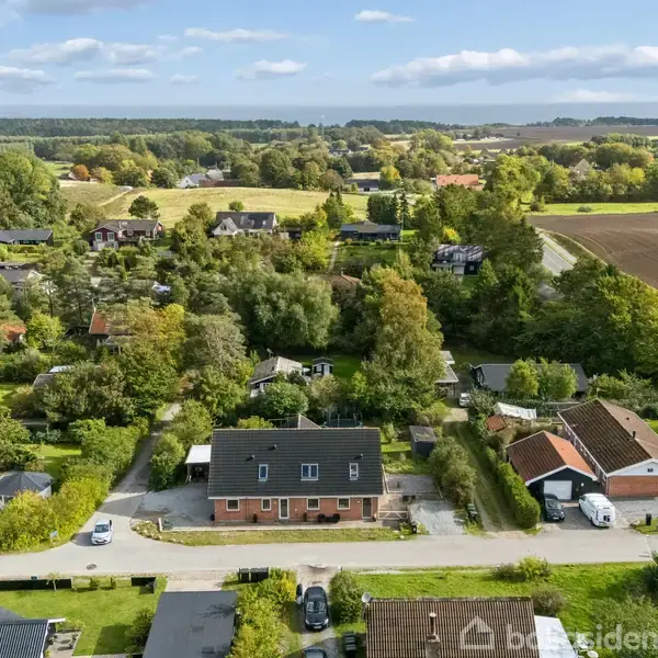 Et rødt murstenshus med sort tag står stille på en villavej omgivet af træer og marker i et landligt område.