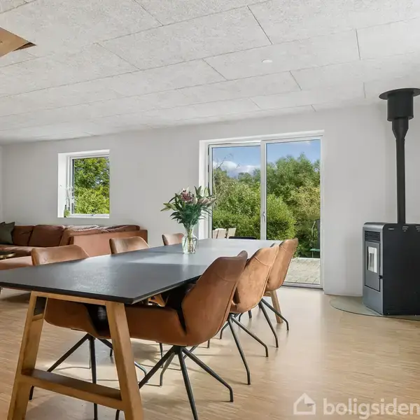 En moderne spisestue med et langt bord og brune stole. Rummet er lyst med store vinduer, en brændeovn i hjørnet og en sofa i baggrunden.