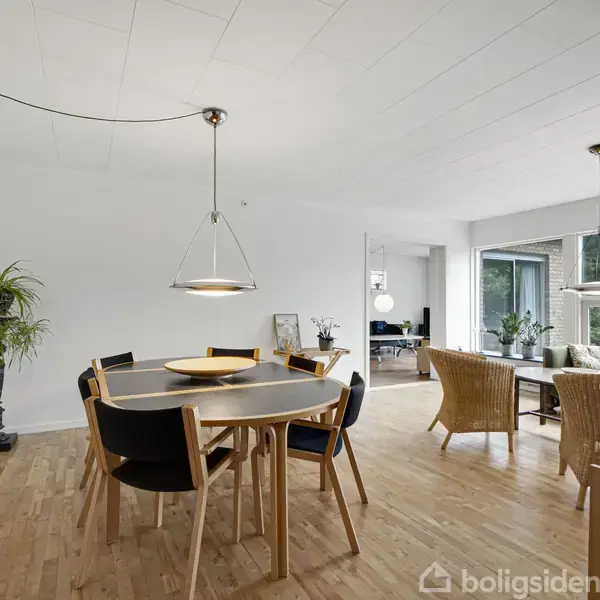 Rundt spisebord med stole står i et lyst rum med trægulve. Store vinduer slipper naturligt lys ind, og en plante står i hjørnet. En stue er i baggrunden.