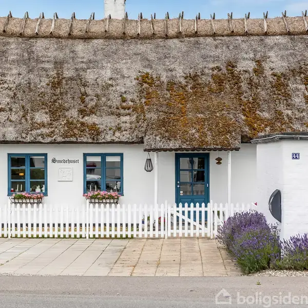 Bindingsværkshus med stråtag, blåmalede vinduer og blomsterkasser. Skilt ved døren viser teksten "Strandhuset". Hvidt stakit foran, omgivet af lavendelbuske. Adresse "43" på muren.