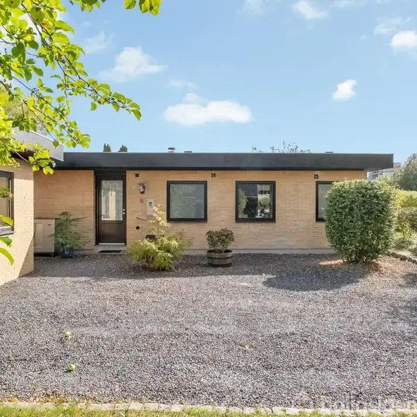 En gulstensbungalow står foran en grusbelagt forplads med planter i krukker, omgivet af grøn vegetation under en klar himmel.
