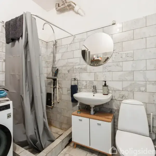 Badeværelse med en brusekabine til venstre ved siden af en vaskemaskine. En håndvask med spejl og skab er monteret på væggen, med et toilet til højre. Vægge er flisebelagte.