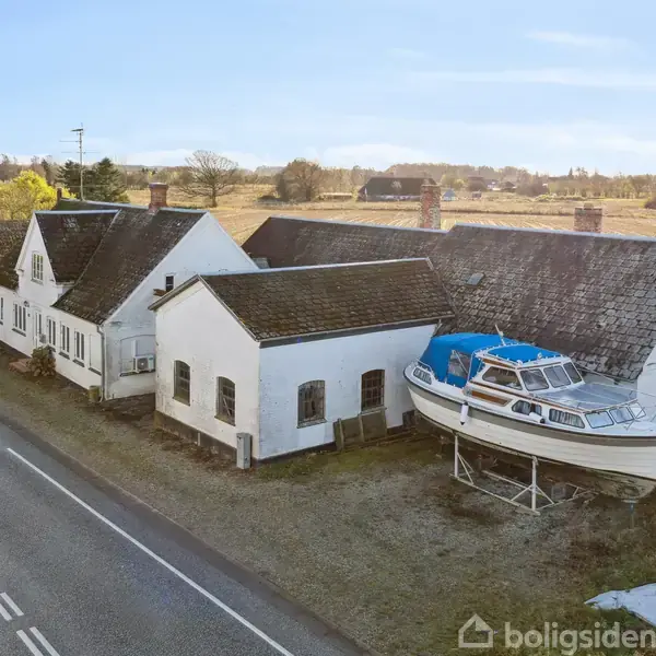 En hvid bygning står ved siden af en vej, med en båd placeret på bukke ved siden af den. Bygningen ligger i et landligt område med marker i baggrunden.