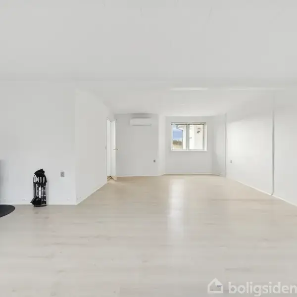 En moderne brændeovn står i hjørnet og varmer det lyse, tomme rum med trægulve og hvide vægge. Et vindue tilføjer naturligt lys til det minimalistiske interiør.