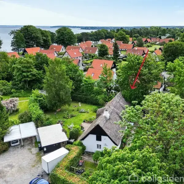 Hus med stråtag omgivet af grønne træer, beliggende i et kvarter med rødt tegltag. En pil peger mod huset, beliggende tæt på kystlinje og udsigt til vand.