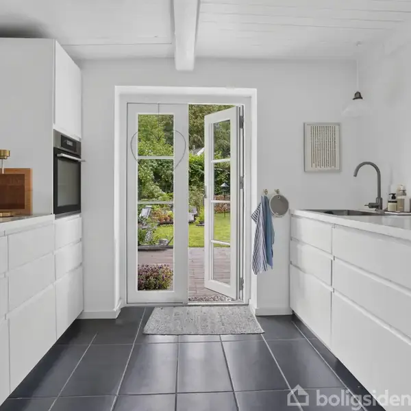 Hvidt moderne køkken med åbne dobbeltdøre fører ud til en have. Der er sort gulvfliser, hvide skabe på begge sider, og en vask med armatur til højre.