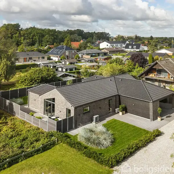 Et moderne enfamiliehus i mursten står omkranset af en velholdt have med græsplæne og buske. Det ligger i et boligområde omgivet af andre huse og grønne træer.