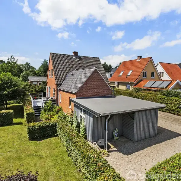 Rødt murstenshus med grå garage ved siden af, omgivet af velplejede hække og græsplæne, stille villakvarter med flere lignende huse i baggrunden under en klar, blå himmel.
