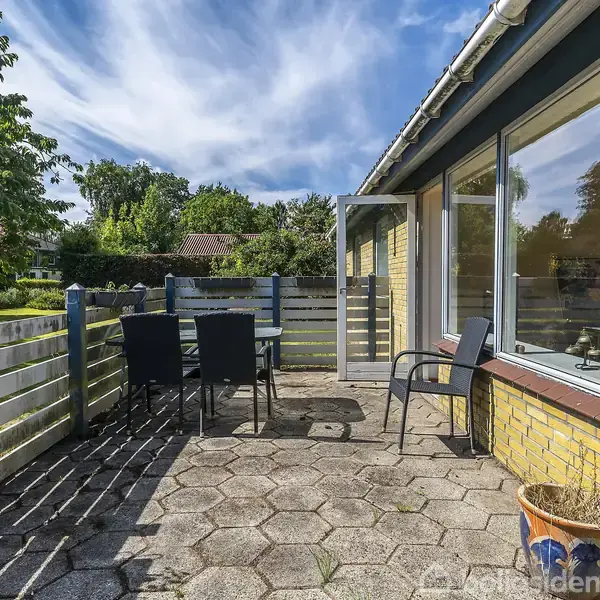 En terrasse med flisebelægning har et bord og fire sorte stole. Terrassen er omgivet af et lavt hegn og en grøn have med træer under en blå himmel.