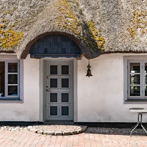 Stråtækt hus med hvid facade og blåmalet dør. Foran står et bord og en stol på en stenbelagt gårdsplads, omgivet af murstensbelægning og en udendørs lampe på væggen.