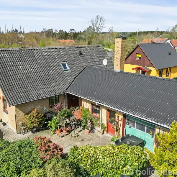 En murstensvilla med sort tag ligger i en velholdt have med buske og blomsterkrukker foran. Omgivelserne indeholder andre huse og en vej, der fører til en mark.