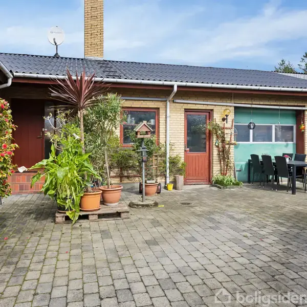 En gårdhave med flisebelægning. Flere blomsterkrukker og planter er placeret rundt omkring. Der er et grillområde med bord og stole ved siden af en garage.