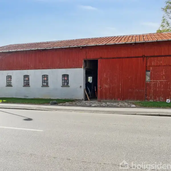 En stor lade med en rød træfacade og små vinduer står ved en vejkant. En åben dør viser interiøret, omgivet af grønne områder og en klar himmel.