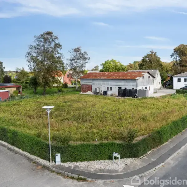 En åben, ubebygget grund med højt græs omkranset af en lav hæk, beliggende ved et vejkryds. I baggrunden ses en gammel bygning og enkelte træer under en blå himmel.