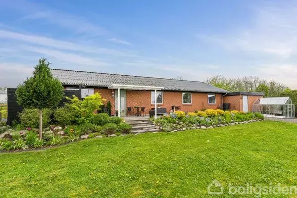 Et rødt murstenshus med sort tag står i en have med en grøn græsplæne og blomsterbede. Et lille drivhus ses i baggrunden under en blå himmel.