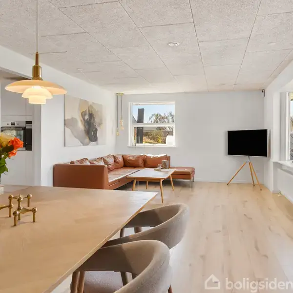 En moderne stue med træbord og stole, dekoreret med blomster i vase. En hjørnesofa og fjernsyn står foran en stor vinduesvæg, der giver naturligt lys og udsigt.