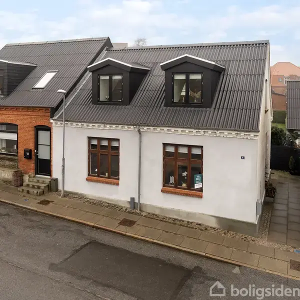 To sammenhængende huse med skrå tage og tagvinduer står på en byvej. Det venstre hus er rødt mursten, det højre er hvidt pudset. Fortovet og gaden er i forgrunden.