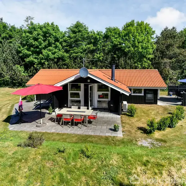 Sommerhus står stille i en grøn have, omgivet af træer. Udendørs spiseområde har rød parasol og stole. Terrasse mod højre har en blå parasol over et sæt havemøbler.