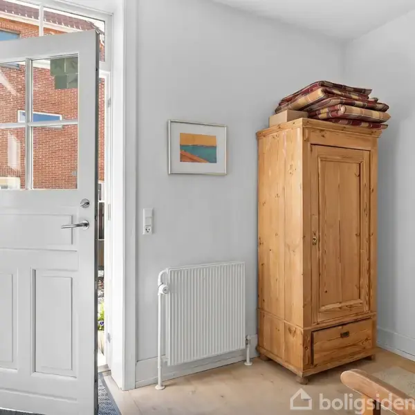 En åben hvid dør i et lyst værelse med et træskab til højre, tæpper ovenpå, ved siden af en radiator og et billede på væggen.