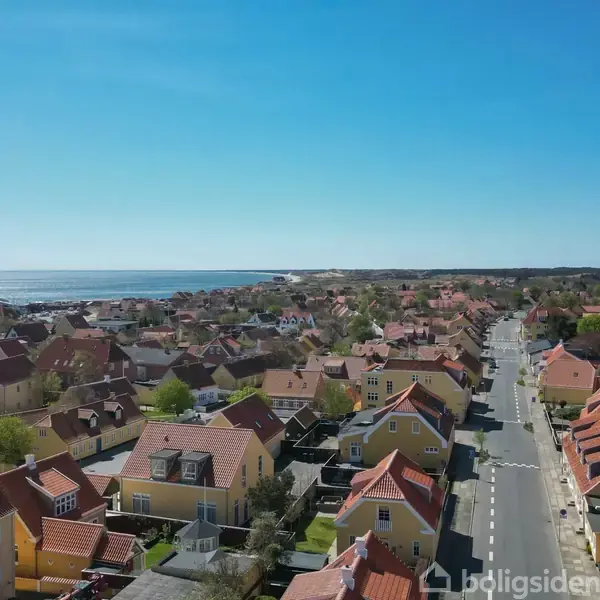 Række af charmerende huse med røde tage strækker sig langs en vej i en solrig kystby. Haven og havet er synlige i baggrunden under en klar blå himmel.