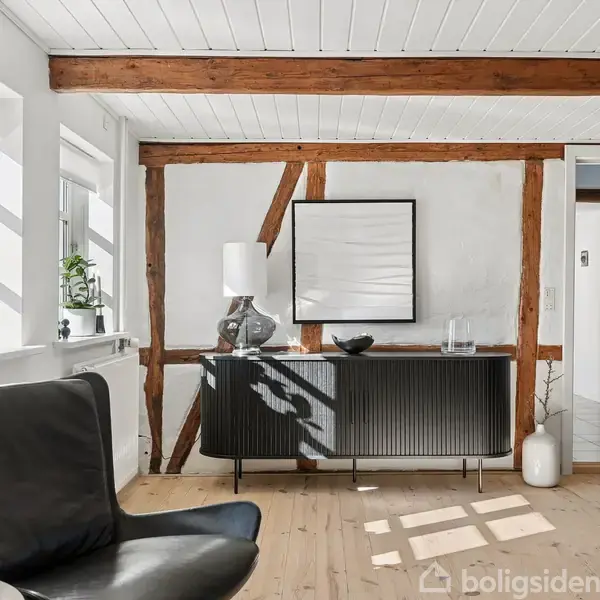 Spisebord med sorte stole, oplyst af sorte lamper, placeret i lys stue med skråloft og ovenlysvindue. Glassofabord og moderne kunst pryder rummet. Dobbelt dør fører til tilstødende rum.