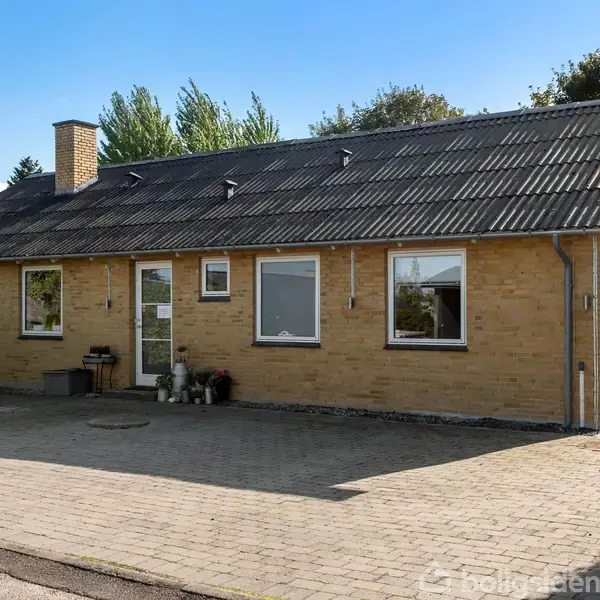 En etplans villa med gul murstensfacade og sort tagstenstag. Foran huset er en flisebelagt indkørsel. Omkring huset er der træer og en klar blå himmel.