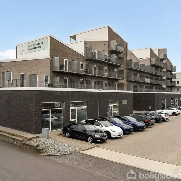 En fler-etagers bygning står med parkerede biler foran. Bygningen har firkantede balkoner. Skiltet på bygningen lyder: "Din sikkerhed - en del af hverdagen."