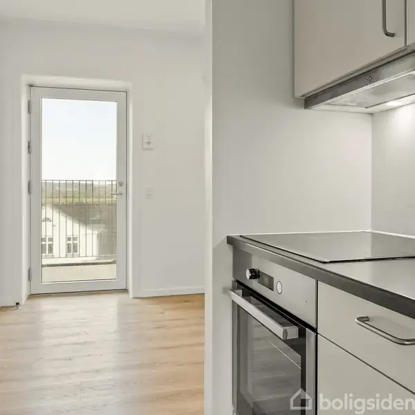 Et moderne køkken med hvide skabe og sort bordplade, hvor ovnen er integreret. Der er udgang til en balkon via en glasdør, og trægulvet strækker sig ind i rummet.
