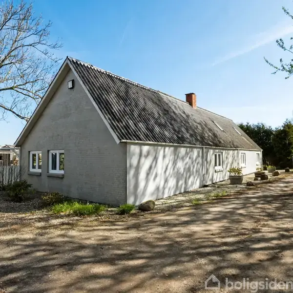Et langstrakt hvidt hus står med en grusvej ved siden af, omgivet af hække og træer under en blå himmel.