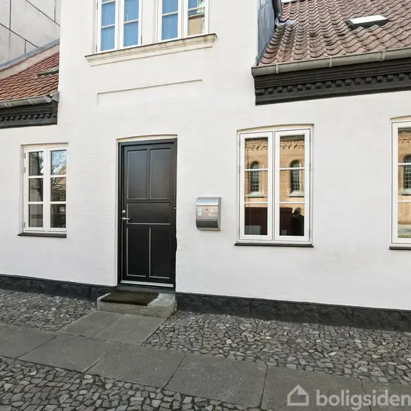 Hvid etageejendom med rødt tegltag og sorte detaljer, står roligt ved en brostensbelagt gade. Otte vinduer og en sort dør giver symmetri og charme til facaden.