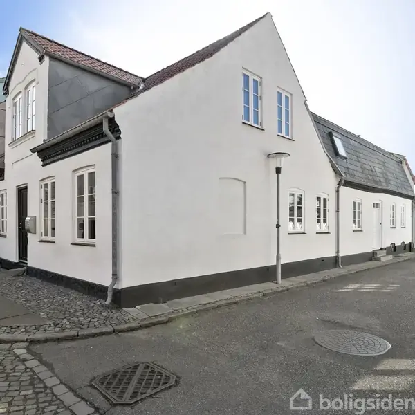 Hvidt hus med skråt tag står i et hjørne af en brostensbelagt gade. På fortovet står en cykel. I baggrunden ses flere bygninger under en blå himmel.