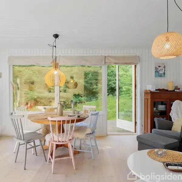 Pejs varmer stue med rundt træbord og stole foran stort vindue mod have. Sofa og lænestol står ved siden af et skab. Lyse træ- og fletlamper hænger fra loftet.
