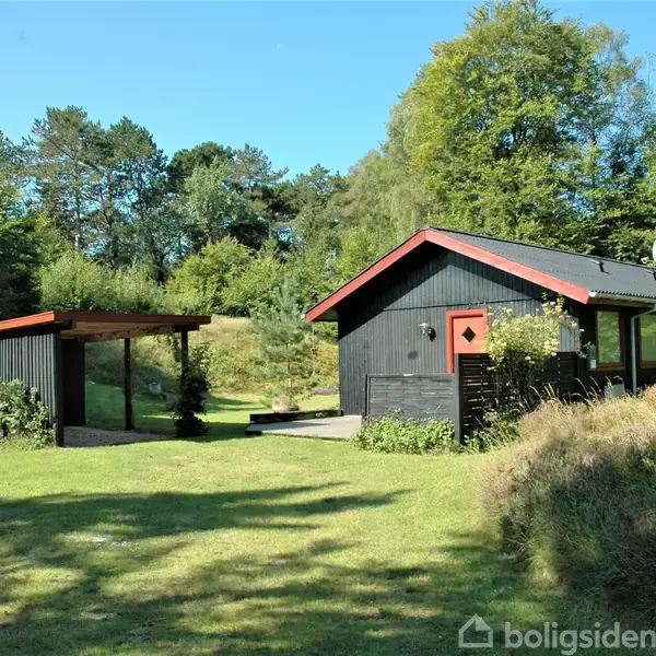 Sort træhus med røde tagkanter står på en frodig græsplæne omgivet af træer. Til venstre er en carport dækket af buske. Sunny, landlig atmosfære.