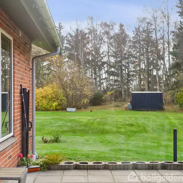 Rødt murstenshus står stille på en græsplæne omgivet af træer, med en bænk foran og en bil parkeret til venstre. Himlen er blå med lette skyer.