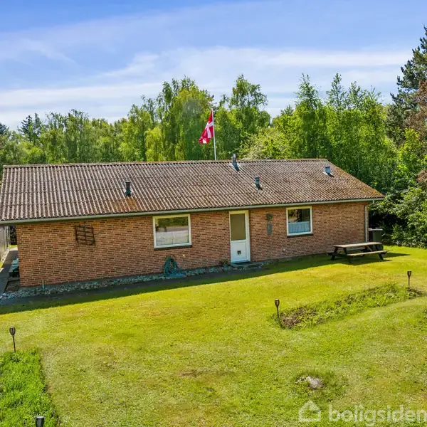 Et rødt murstenshus med terrasse står på en grøn græsplæne omgivet af træer. Til venstre ses et flag på en flagstang.