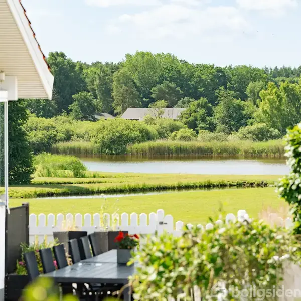 Stue med store vinduer åbner mod en terrasse med udsigt over en grøn have. Planter på vindueskarmene skaber en naturlig atmosfære. Ingen tekst tilgængelig.