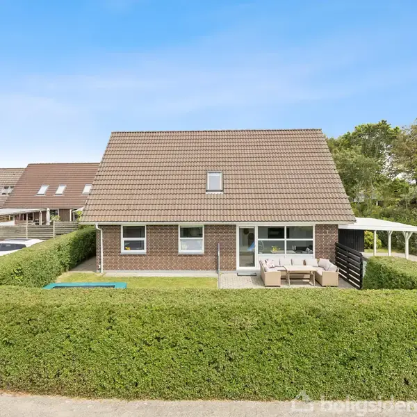Et murstenshus med saddeltag omgivet af tætklippet hæk. Huset har en terrasse med møbler. I baggrunden ses flere andre huse og tætte træer. Ingen tekst er synlig.