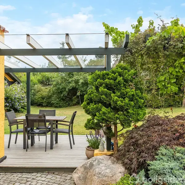 Terrassen har et spisebord med stole, der står under en glasoverdækning. Omkring terrassen er der frodig beplantning og græsplæne ved siden af et gult hus.