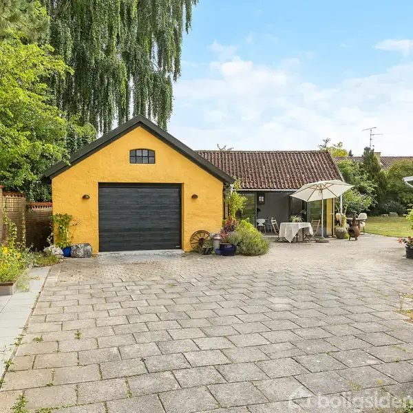 Et gult hus med gråt tag står stille omgivet af grøn beplantning. Foran huset er en flisebelagt indkørsel og en terrasse med parasol og havemøbler.