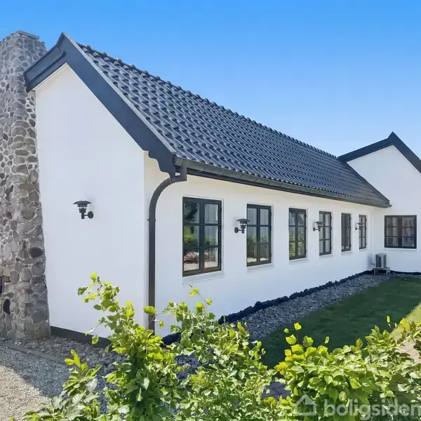 Hvidt hus med sorte tagsten og stor skorsten står i en velholdt have. Flere vinduer langs væggen giver lys. Grusbelagt sti omkranser huset, omgivet af grønne planter.