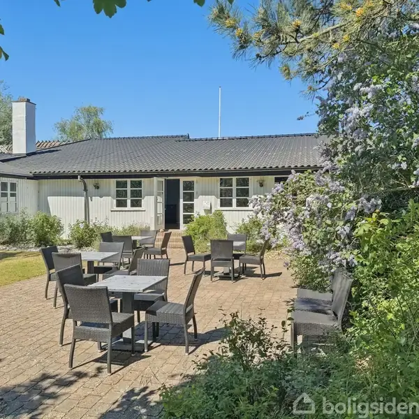 Hvid bygning med sort tag står stille omgivet af frodig have og blå himmel. I forgrunden er der flere udendørs siddepladser på en terrasse med flisebelægning.