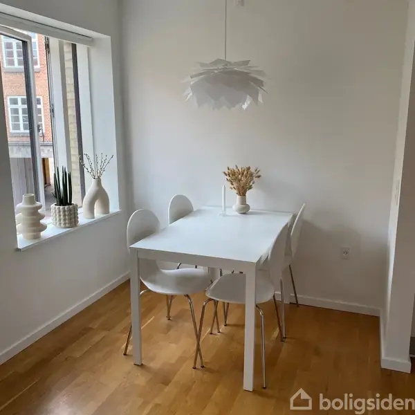 Et hvidt spisebord med fire hvide stole står i et hjørne af et lyst rum. På bordet er der en vase med tørrede blomster. Vinduet har keramik og planter.