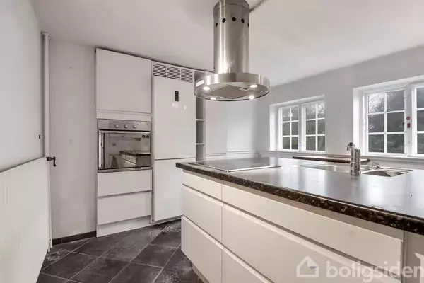 Et moderne køkken med hvidt køleskab, indbygningsovn og emhætte over en køkkenø. Flisegulv og store vinduer giver lys og en stilfuld atmosfære.