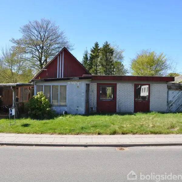 Et lille hus med rødt tag og hvide vægge står ubenyttet. Det er omgivet af en græsplæne og har en carport på venstre side. Det er en solrig dag.